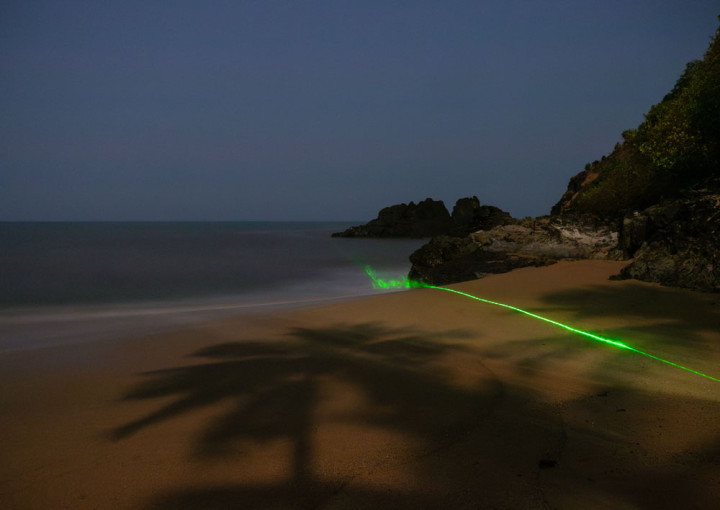 The photo shows the sea at night in the background, rocks can be seen on the right and a sandy beach in the foreground with a neon-green beam of light running across it