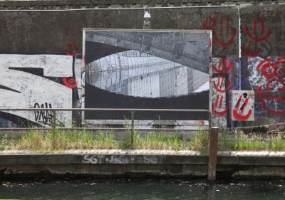 Die Fotografie, aufgezogen als Plakatwand an der Brückenmauer, zeigt die Spiegelung der Mühlendammbrücke, die sich mit der realen Brückenstruktur zu einer Form verbindet, die an ein Auge erinnert
