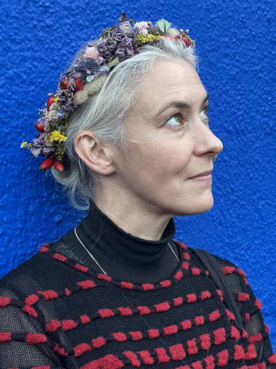 Die Künstlerin Alex Lebus steht vor einer blauen Wand und trägt einen Blumenkranz