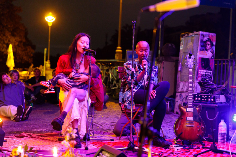 Zwei Künstler*innen der Jamsession singen. Die eine Sängerin stillt währenddessen ihr Baby an ihrer Brust.